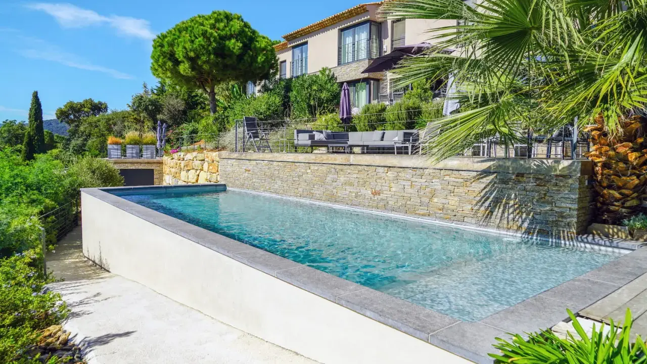 Belle piscine semi enterrée avec un habillage en pierre naturelle, entourée de verdure luxuriante et d'une villa moderne.