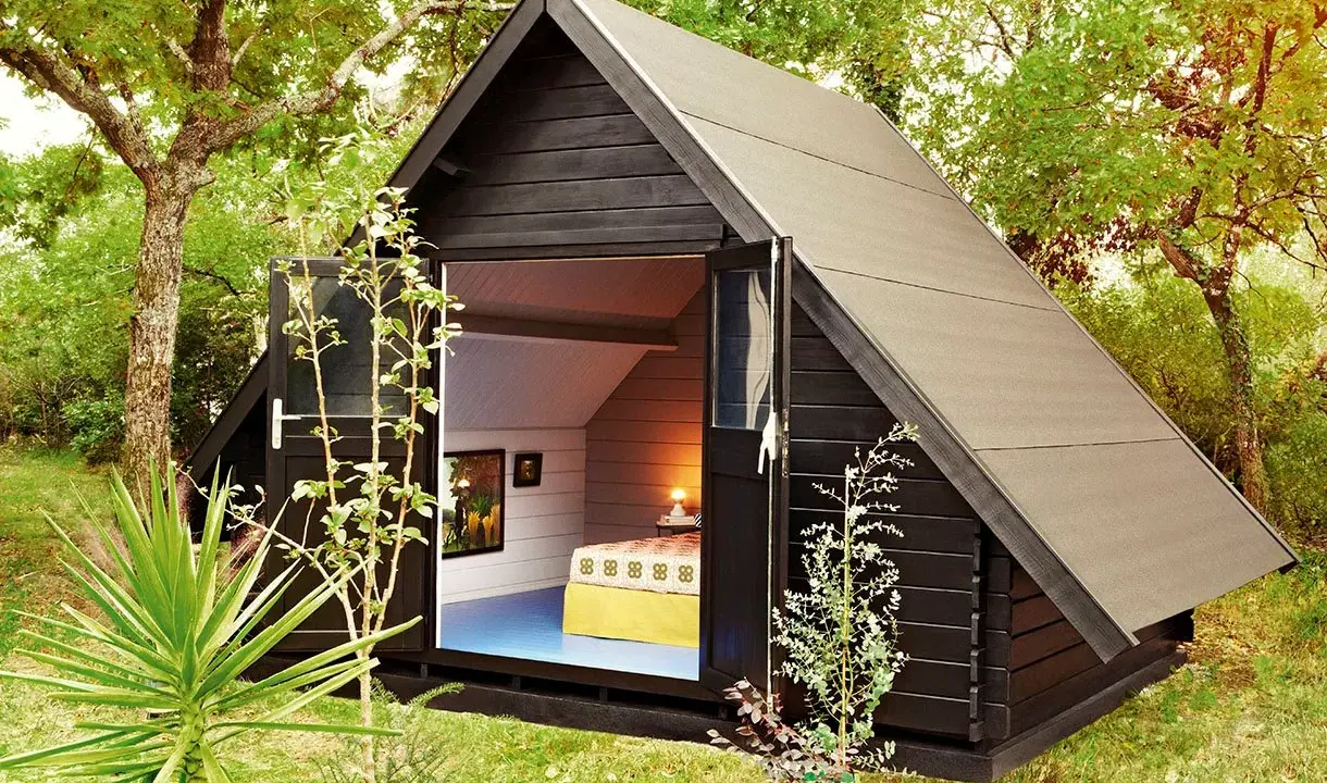 Aménagement abri de jardin : une cabane moderne en bois noir avec une porte ouverte révélant un lit douillet et des tableaux.