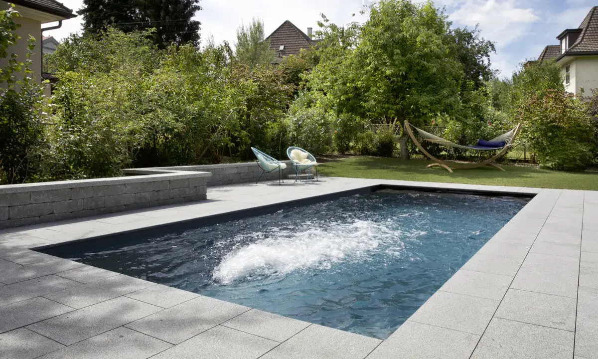Aménagement paysager d'une piscine creusée avec terrasse en pierre, deux chaises bleues et un hamac invitant à la détente.