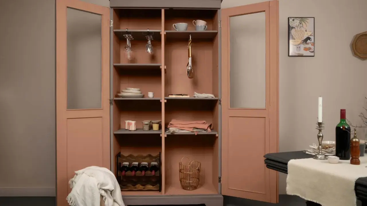 Armoire vitrée avec un relooking meuble 2 couleurs : gris anthracite et rose poudré. Elle contient vaisselle, verres, bouteilles de vin et paniers.