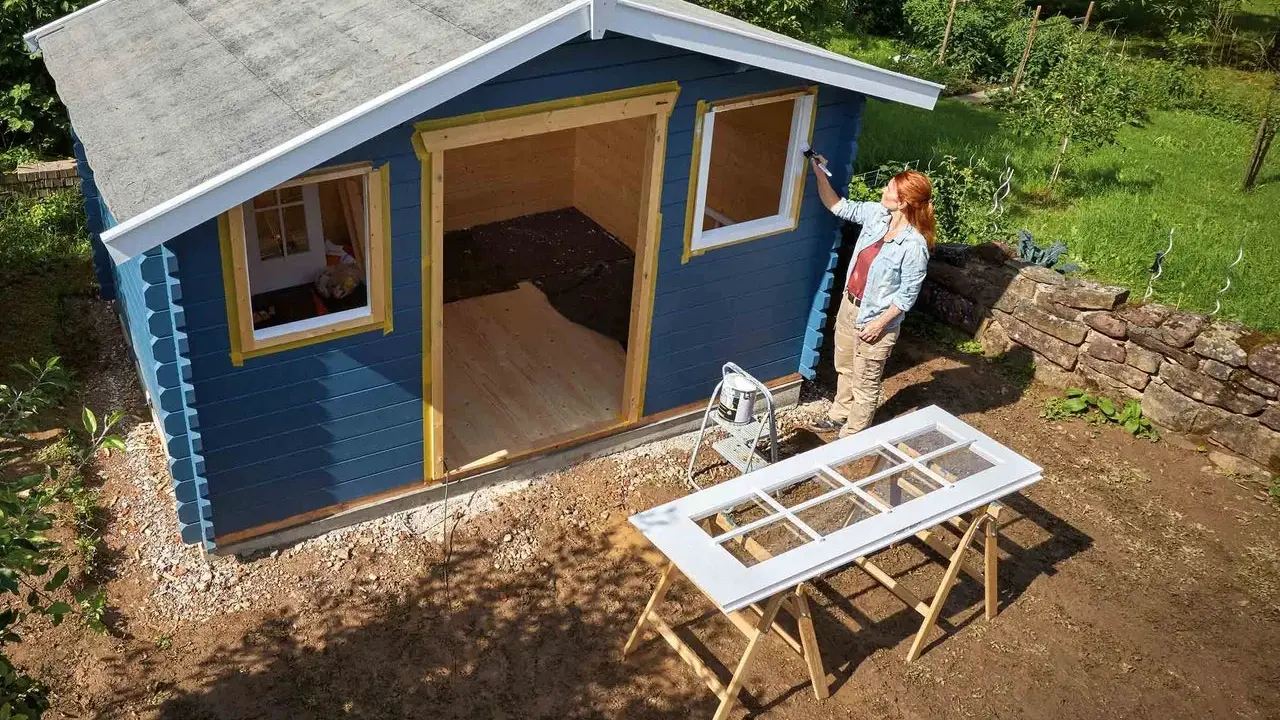 Une femme peint les contours des fenêtres d'une cabane de jardin bleue. Le toit de la cabane est en cours de finition.