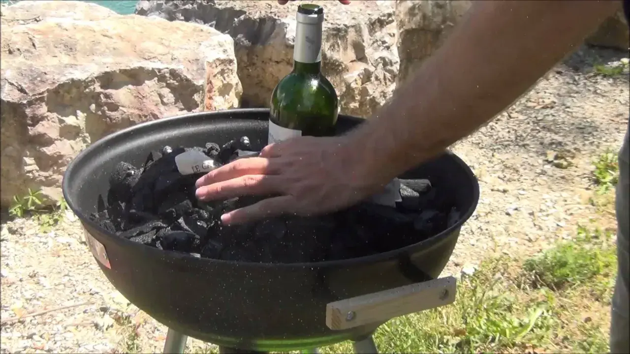 Main d'homme plaçant une bouteille de vin verte sur du charbon de bois dans un barbecue, étape pour comment allumer un barbecue.