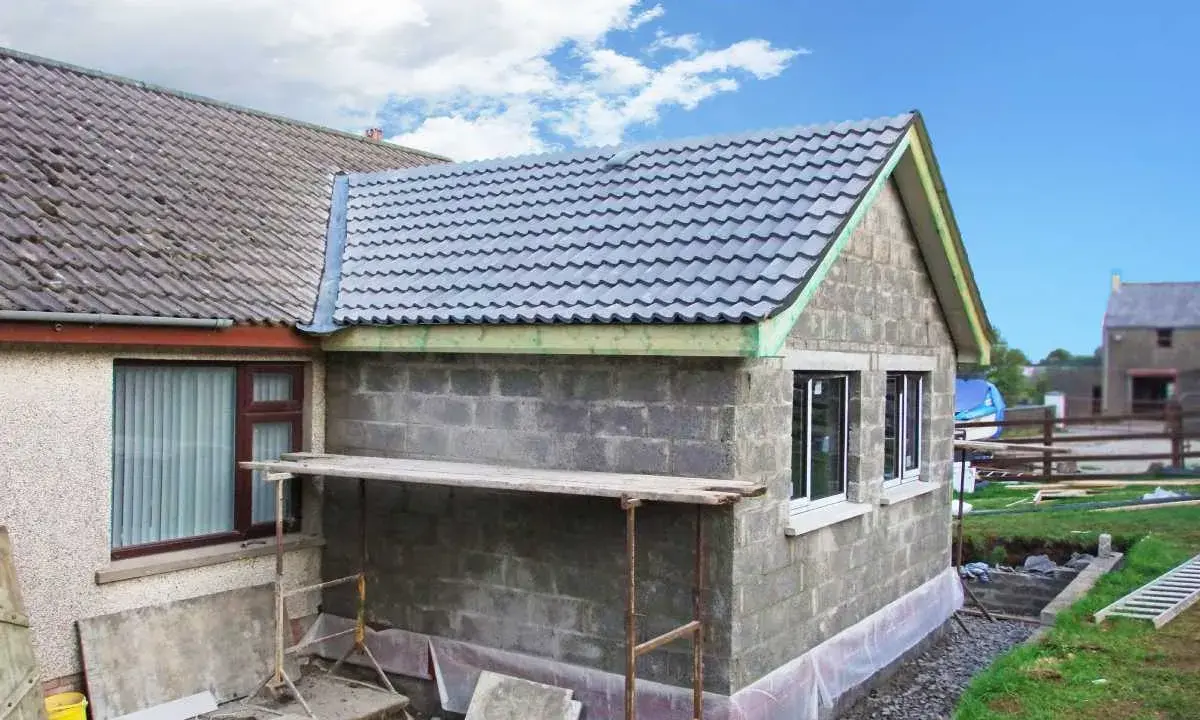 Agrandissement maison sur le côté en cours de construction, murs en parpaings, nouveau toit gris, échafaudage.