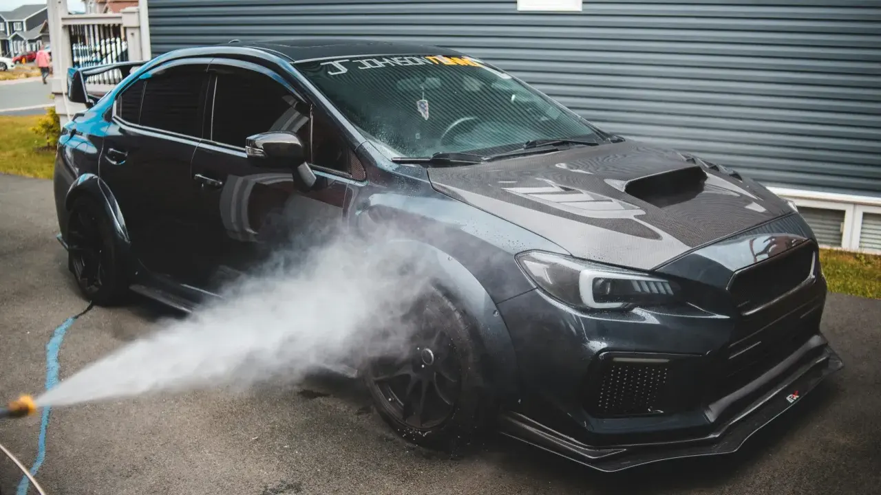Nettoyage d'une voiture de sport sombre. Un jet d'eau est dirigé vers la roue avant, peut-être pour enlever mousse sur goudron vinaigre blanc.