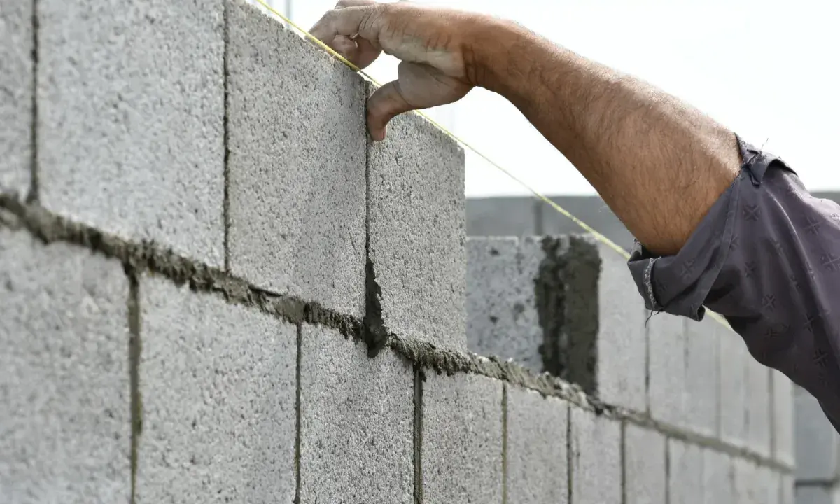 Main d'un maçon plaçant une brique sur un mur en parpaing, guidé par un fil à plomb.