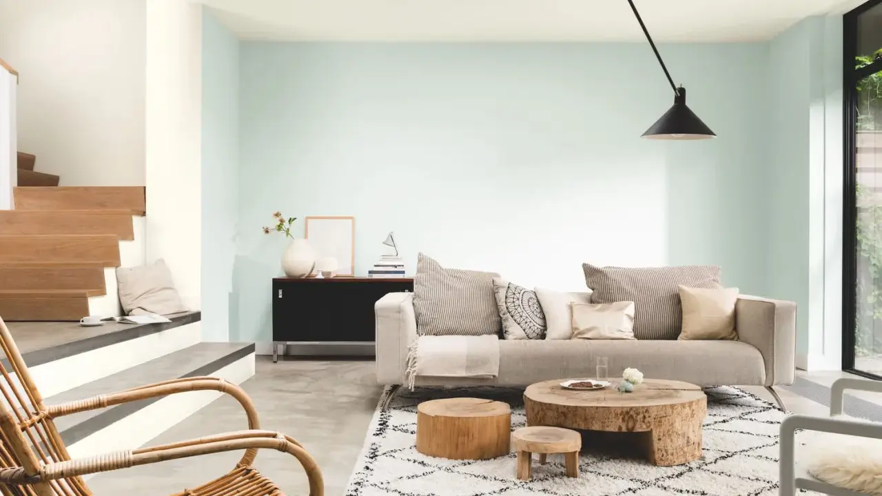 Salon lumineux aux murs couleur vert d'eau, avec un canapé beige, des tables basses en bois brut et un fauteuil en rotin.