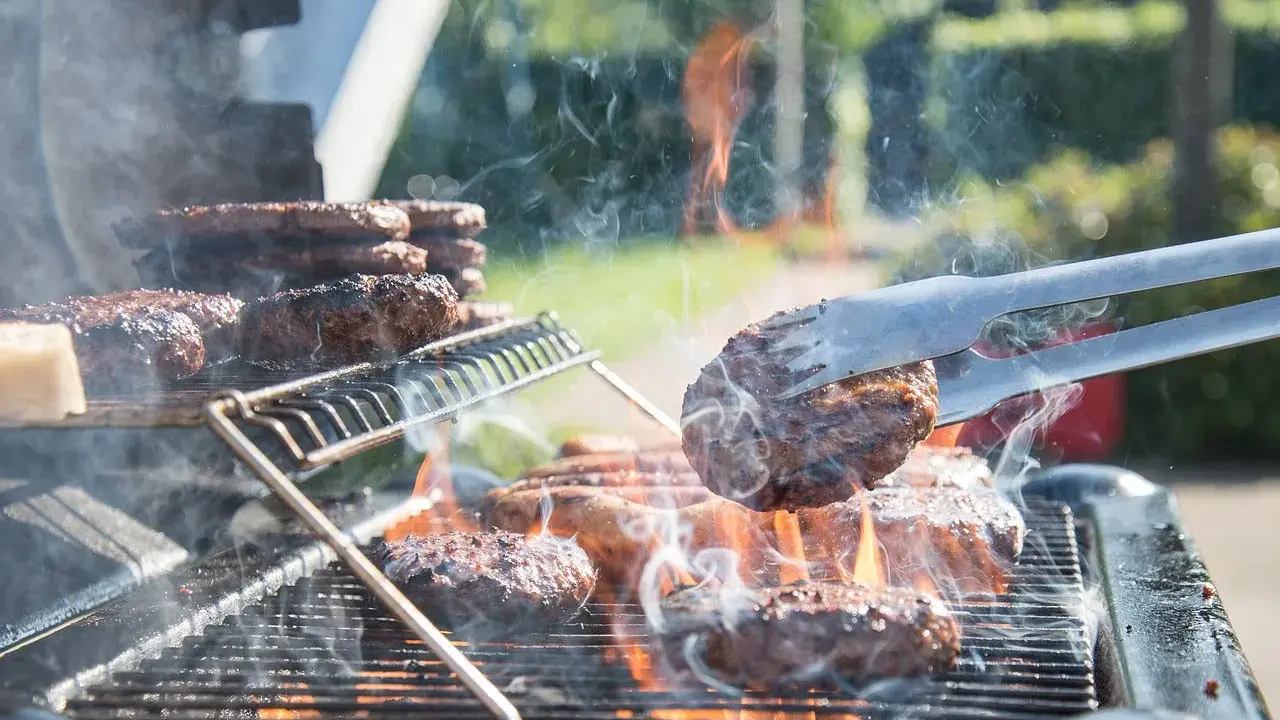 Des hamburgers grillent sur un barbecue à gaz. La cuisson est intense, la fumée s'élève. Nettoyer barbecue gaz après ce festin sera nécessaire.