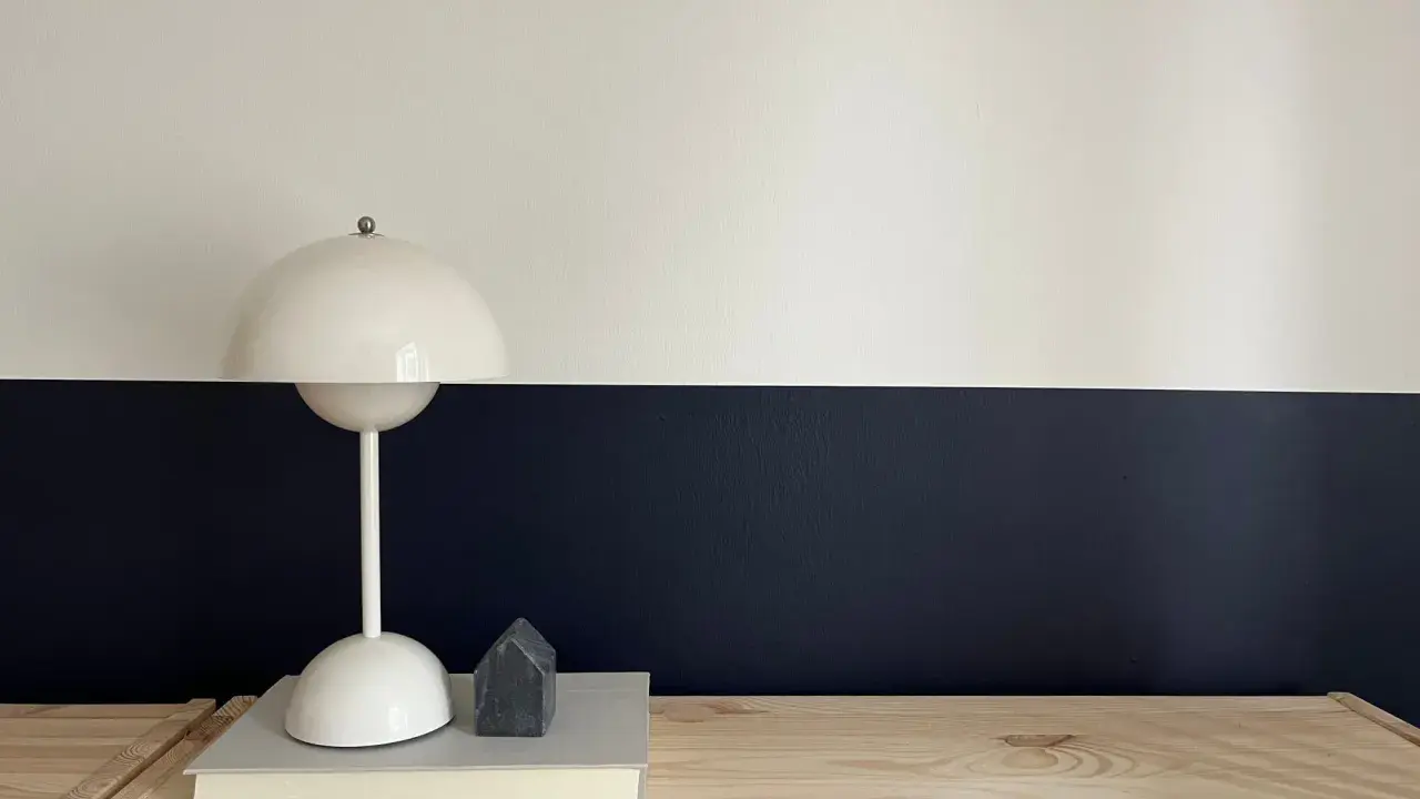Lampe blanche sur un livre, devant un mur deux couleurs différentes, blanc et bleu nuit. Un petit objet gris en forme de maison est posé sur le livre.