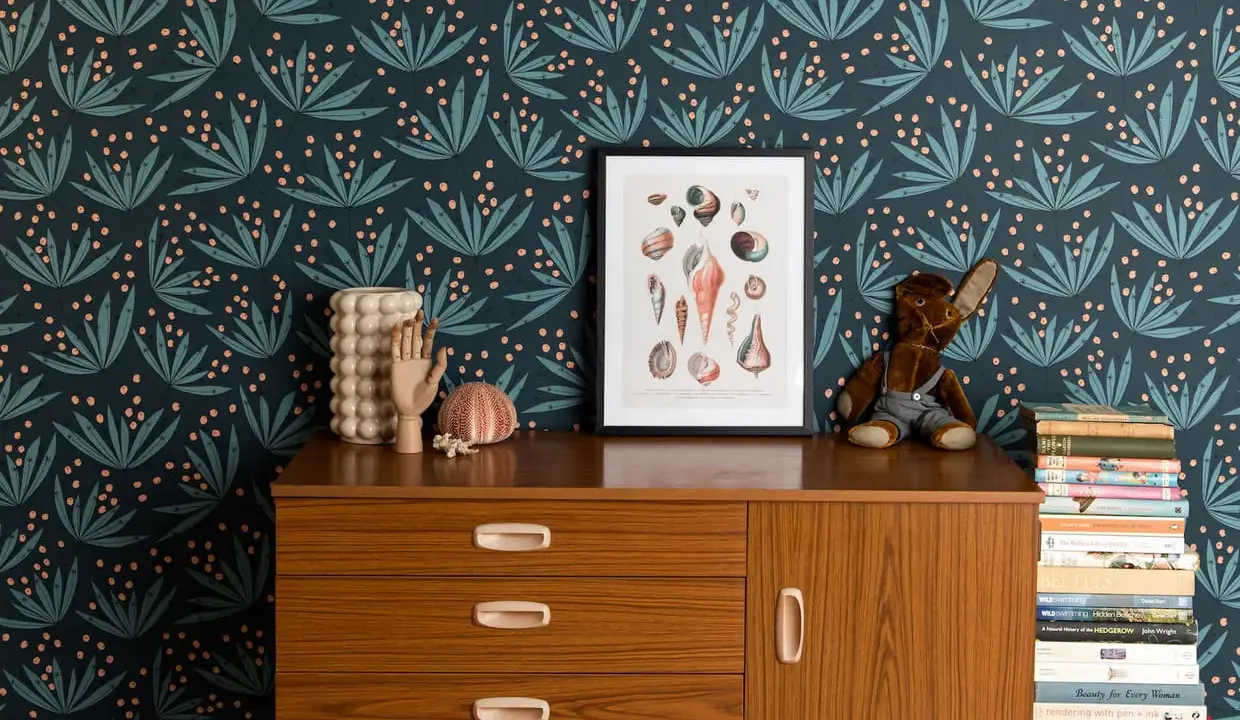 Idée motif peinture mur : papier peint botanique bleu nuit avec des feuilles stylisées et des baies roses. Un cadre de coquillages et une commode vintage complètent la scène.