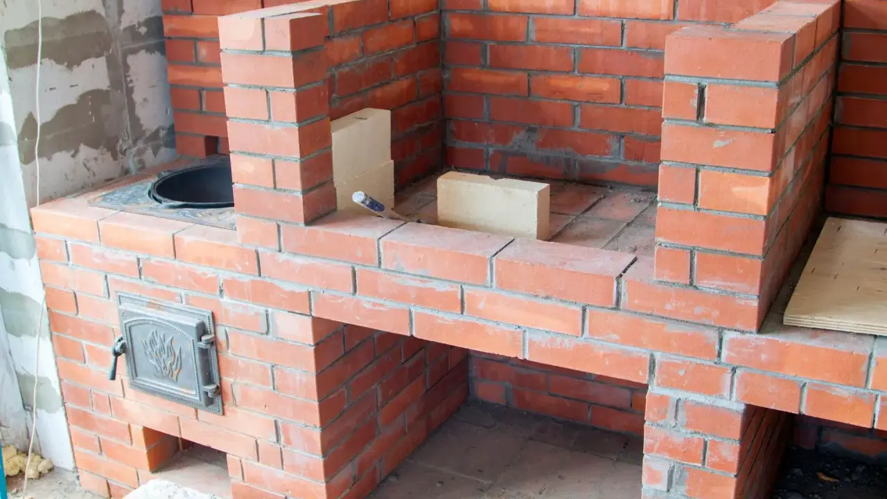 Construction d'un barbecue en brique, avec une porte en métal ornée et des briques claires pour la finition.