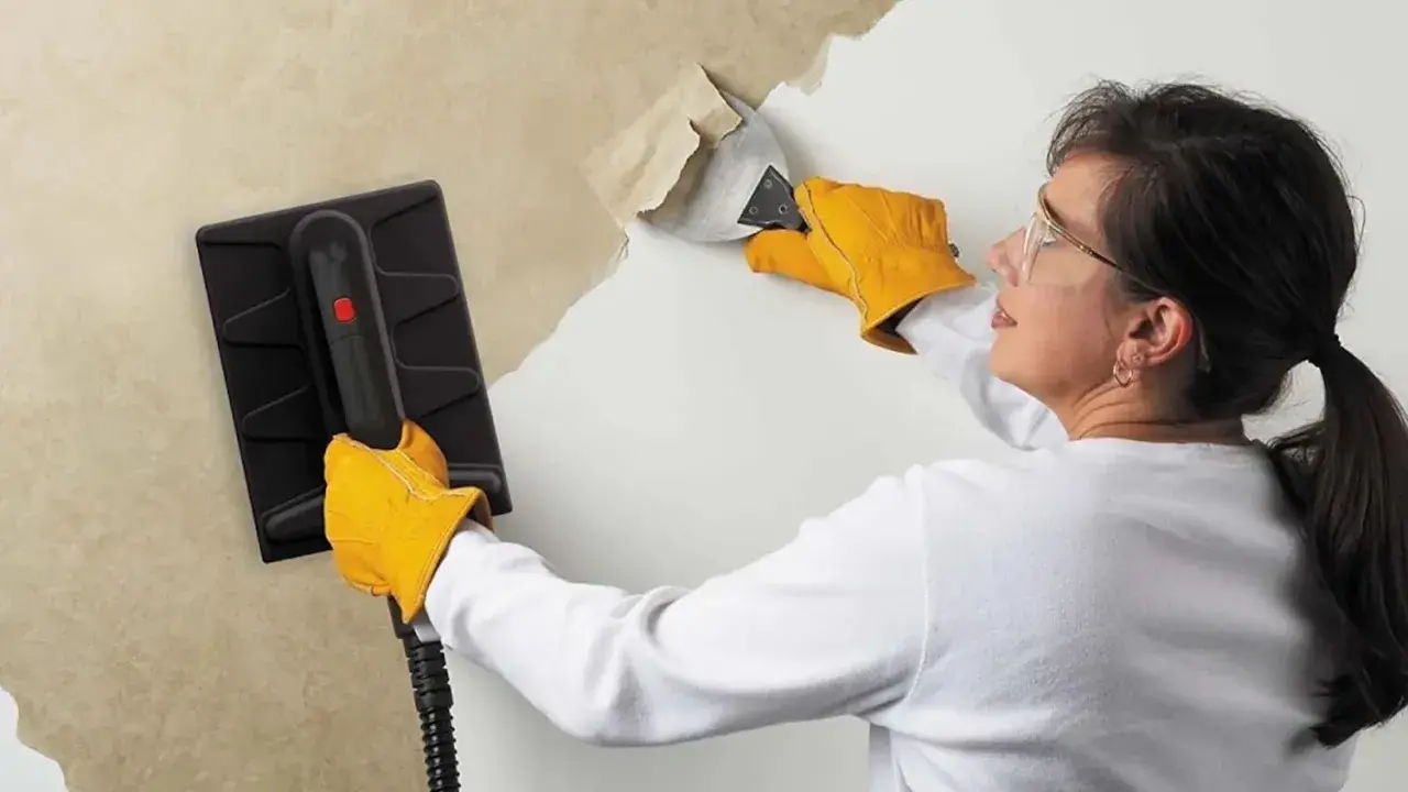 Une femme, portant des gants jaunes, utilise un appareil pour décoller du papier peint ancien d'un mur.