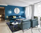Salon moderne aux murs bleu canard, avec canapé velours, fauteuils assortis, tapis graphique et éclairage d'ambiance.