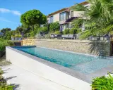Belle piscine semi enterrée avec un habillage en pierre naturelle, entourée de verdure luxuriante et d'une villa moderne.