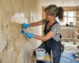 Une femme en salopette bleue, portant des gants bleus et des lunettes, utilise une éponge et une brosse pour lessiver un mur avant de le peindre.