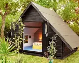 Aménagement abri de jardin : une cabane moderne en bois noir avec une porte ouverte révélant un lit douillet et des tableaux.