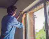 Un homme ajuste le joint d'une nouvelle pose fenêtre PVC. Le soleil brille à travers la vitre.