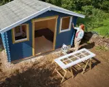 Une femme peint les contours des fenêtres d'une cabane de jardin bleue. Le toit de la cabane est en cours de finition.