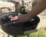 Main d'homme plaçant une bouteille de vin verte sur du charbon de bois dans un barbecue, étape pour comment allumer un barbecue.