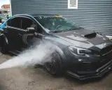 Nettoyage d'une voiture de sport sombre. Un jet d'eau est dirigé vers la roue avant, peut-être pour enlever mousse sur goudron vinaigre blanc.