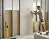 Un homme visse une plaque de placo sur une structure métallique. Des outils et des matériaux de construction sont visibles.