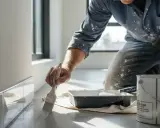 Un peintre applique une couche de peinture sur une plinthe, juste au-dessus du carrelage.