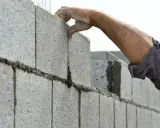 Main d'un maçon plaçant une brique sur un mur en parpaing, guidé par un fil à plomb.