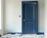Peinture porte intérieure bleu marine, avec des touches de bleu clair sur le mur. Des rouleaux et pinceaux sont au sol, prêts pour la prochaine étape.