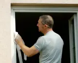 Homme en cours de pose de fenêtre en rénovation, nettoyant le cadre avec un chiffon. Un crayon est coincé derrière son oreille.