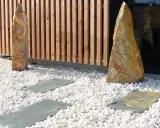 Deux pierres dressées sur une allée en gravier blanc, bordée de dalles grises et d'une palissade en bois.