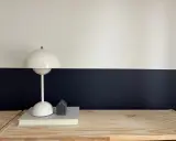 Lampe blanche sur un livre, devant un mur deux couleurs différentes, blanc et bleu nuit. Un petit objet gris en forme de maison est posé sur le livre.