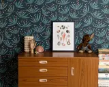 Idée motif peinture mur : papier peint botanique bleu nuit avec des feuilles stylisées et des baies roses. Un cadre de coquillages et une commode vintage complètent la scène.