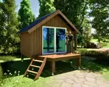 Un homme porte sa fille sur la terrasse d'un abri de jardin sur plot, entouré de verdure.