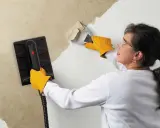 Une femme, portant des gants jaunes, utilise un appareil pour décoller du papier peint ancien d'un mur.