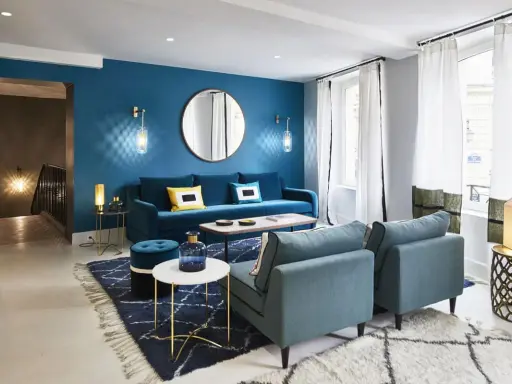 Salon moderne aux murs bleu canard, avec canapé velours, fauteuils assortis, tapis graphique et éclairage d'ambiance.