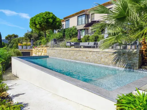 Belle piscine semi enterrée avec un habillage en pierre naturelle, entourée de verdure luxuriante et d'une villa moderne.