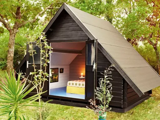 Aménagement abri de jardin : une cabane moderne en bois noir avec une porte ouverte révélant un lit douillet et des tableaux.