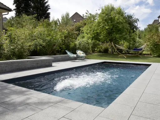 Aménagement paysager d'une piscine creusée avec terrasse en pierre, deux chaises bleues et un hamac invitant à la détente.