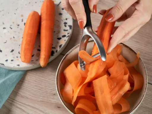 Des carottes épluchées tombent en rubans dans un bol. Une astuce pour comment conserver les carottes : les préparer ainsi pour une cuisson rapide.