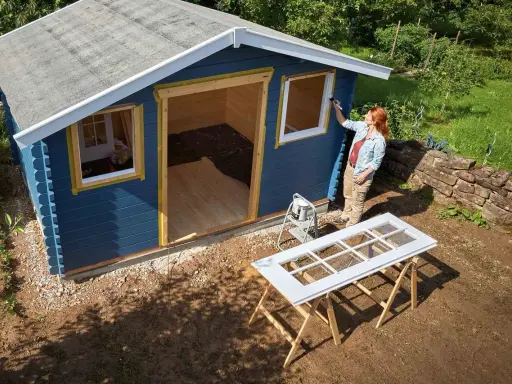 Une femme peint les contours des fenêtres d'une cabane de jardin bleue. Le toit de la cabane est en cours de finition.