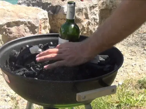 Main d'homme plaçant une bouteille de vin verte sur du charbon de bois dans un barbecue, étape pour comment allumer un barbecue.