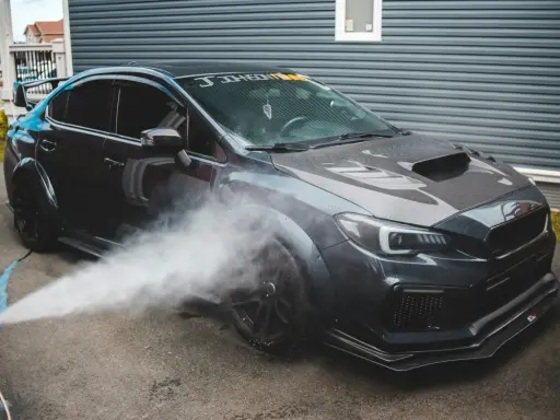 Nettoyage d'une voiture de sport sombre. Un jet d'eau est dirigé vers la roue avant, peut-être pour enlever mousse sur goudron vinaigre blanc.