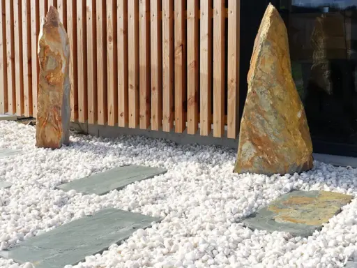Deux pierres dressées sur une allée en gravier blanc, bordée de dalles grises et d'une palissade en bois.