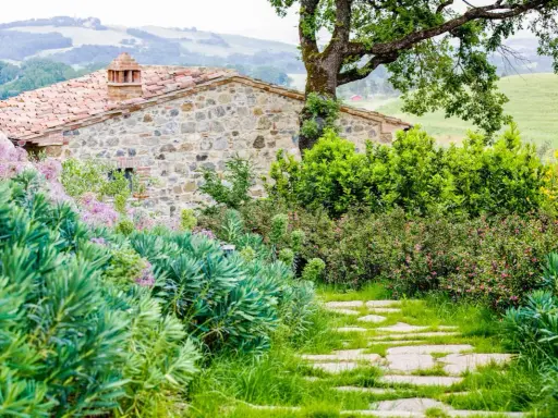 Un chemin de dalles de pierre serpente à travers un jardin luxuriant, bordé de divers types de haies. Des fleurs roses et des arbustes verdoyants entourent une maison en pierre toscane.