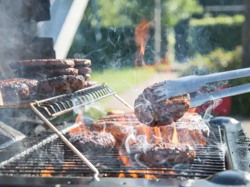 Des hamburgers grillent sur un barbecue à gaz. La cuisson est intense, la fumée s'élève. Nettoyer barbecue gaz après ce festin sera nécessaire.