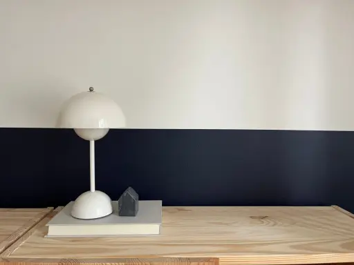Lampe blanche sur un livre, devant un mur deux couleurs différentes, blanc et bleu nuit. Un petit objet gris en forme de maison est posé sur le livre.