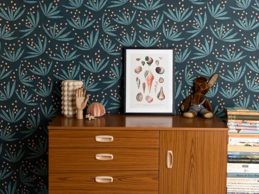Idée motif peinture mur : papier peint botanique bleu nuit avec des feuilles stylisées et des baies roses. Un cadre de coquillages et une commode vintage complètent la scène.
