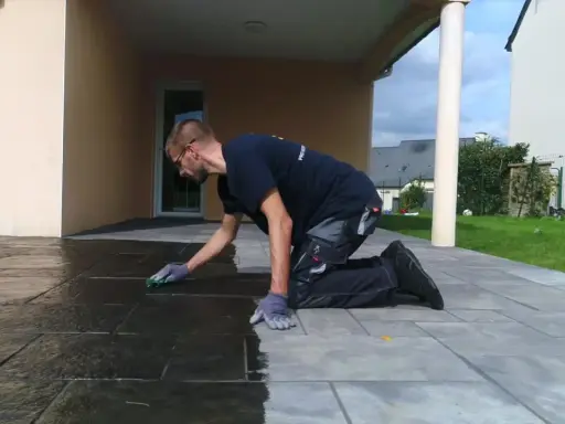 Un homme agenouillé effectue le nettoyage de dalles de terrasse avec une éponge verte, redonnant leur éclat à la pierre.