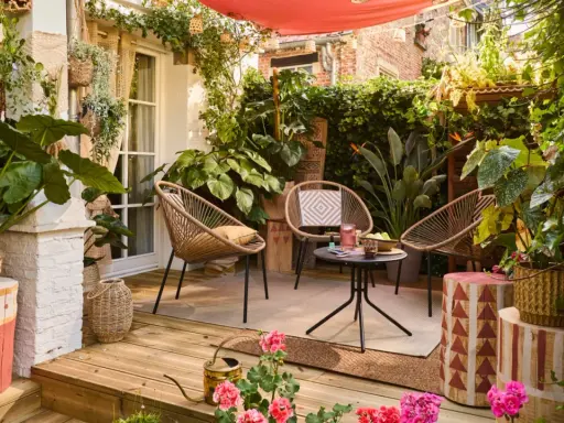 Aménager une petite terrasse sans se ruiner : trois fauteuils en rotin, une table basse, des plantes luxuriantes et une toile rouge créent un coin cosy et abordable.