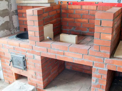 Construction d'un barbecue en brique, avec une porte en métal ornée et des briques claires pour la finition.