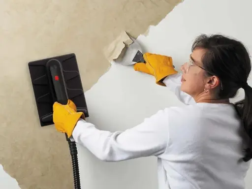 Une femme, portant des gants jaunes, utilise un appareil pour décoller du papier peint ancien d'un mur.